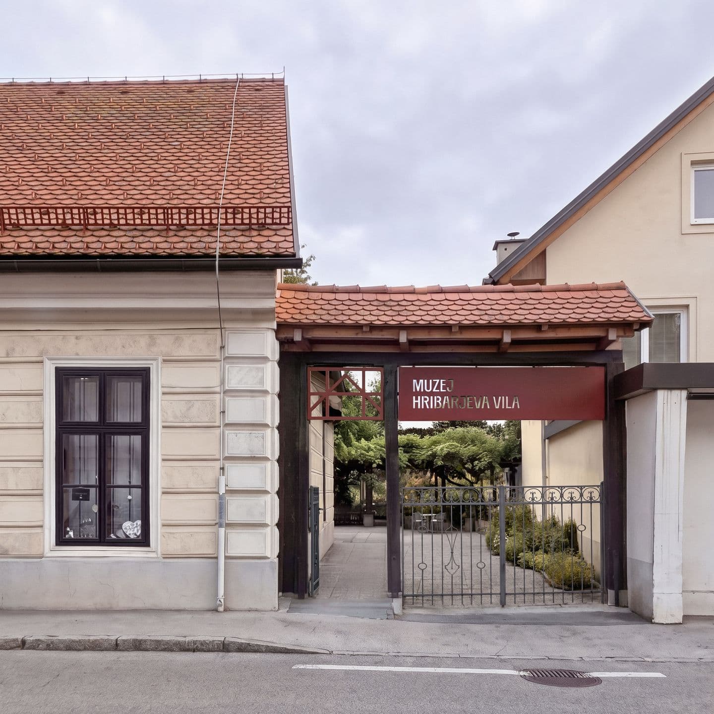 A covered entrance with a sign reading "MUZEJ HRIBARJEVA VILA" and a wrought-iron gate stands between two tile-roofed buildings leading to a courtyard.